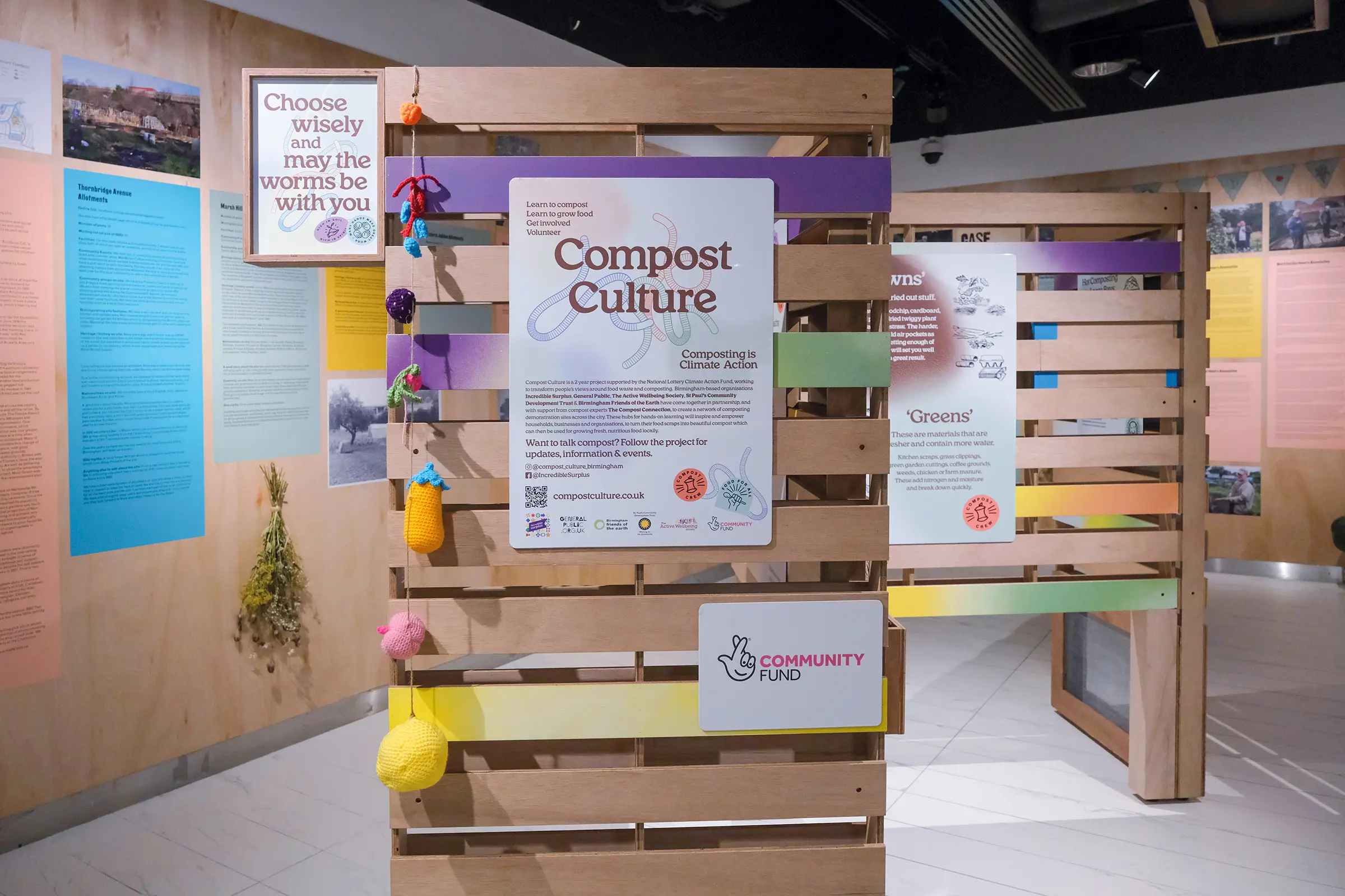 An image of multicolored exhibition panels, in a white gallery space. The panels are made from recycled crates and sections of them are dyed with different colours. In the centre of the main panel is a white board with text reading 'Compost Culture' on it.
