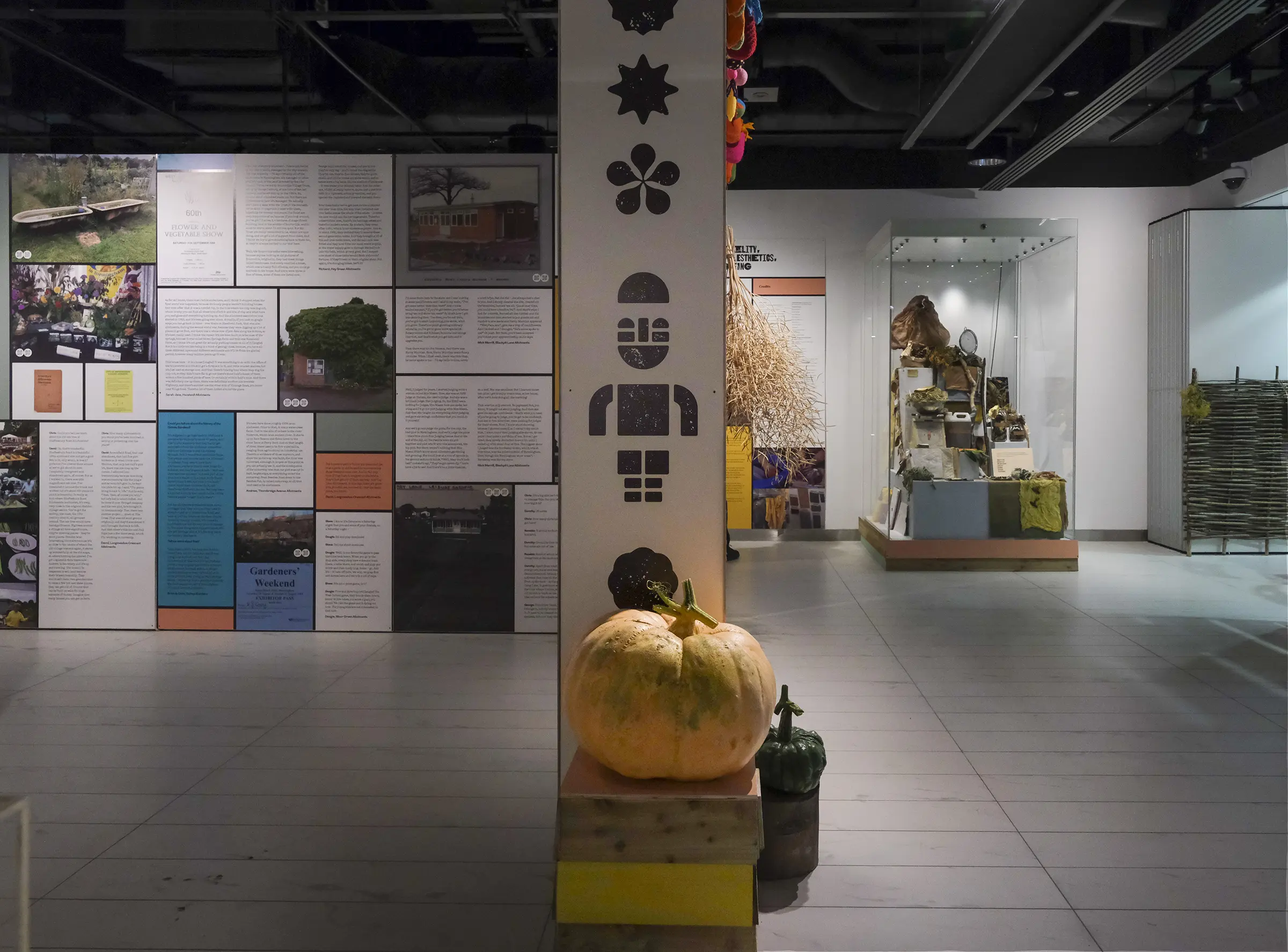 An image of multicolored exhibition panels, in a white gallery space. In the centre of the image is a column with images of figures and plants made from a woodblock print typeface on it. In front of the column is a large orange pumpkin, sat atop a wooden crate.