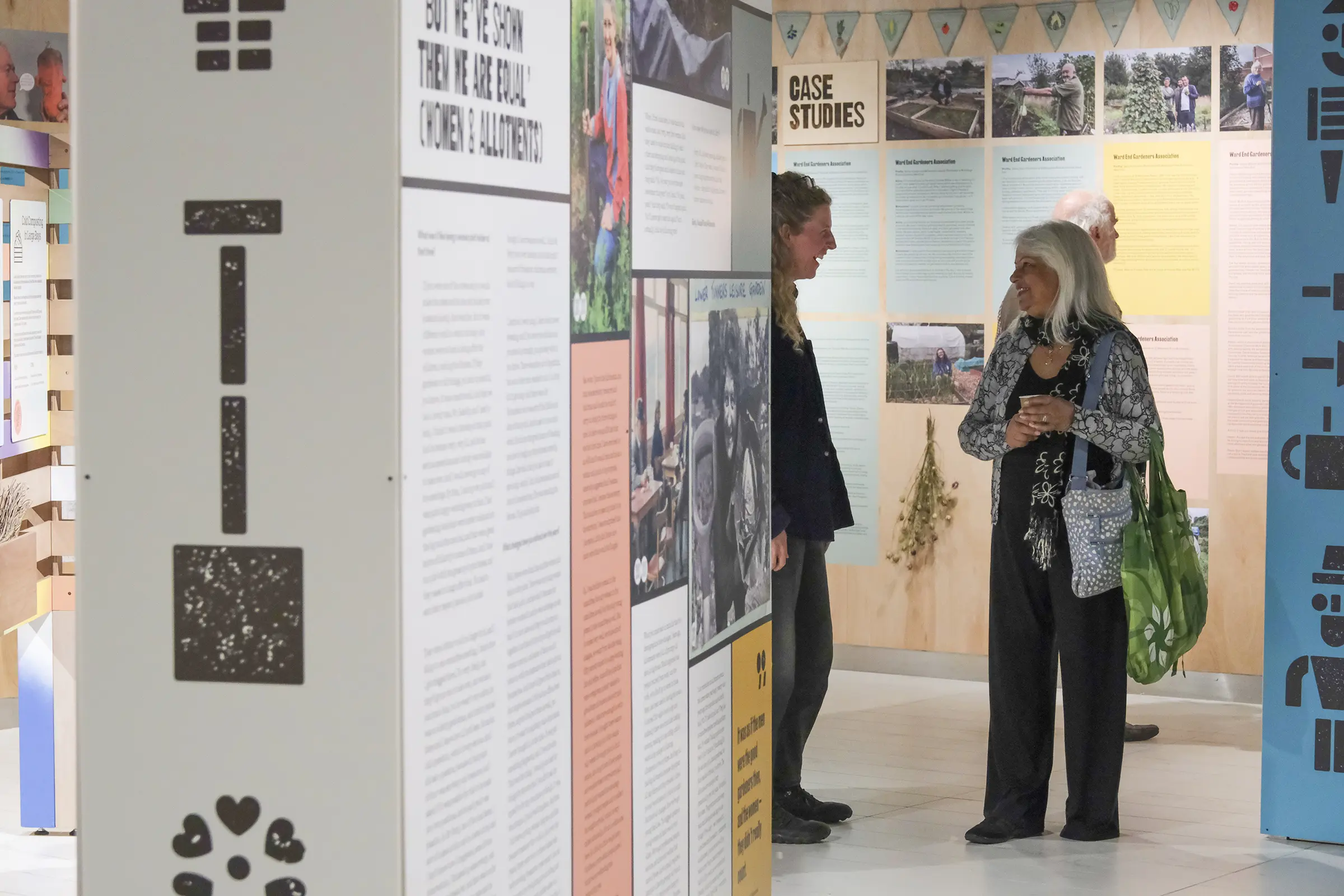 An image of two people talking in the background of an exhibition space. In front of them is an exhibition panel, with an image of a spade made from a woodblock print font.