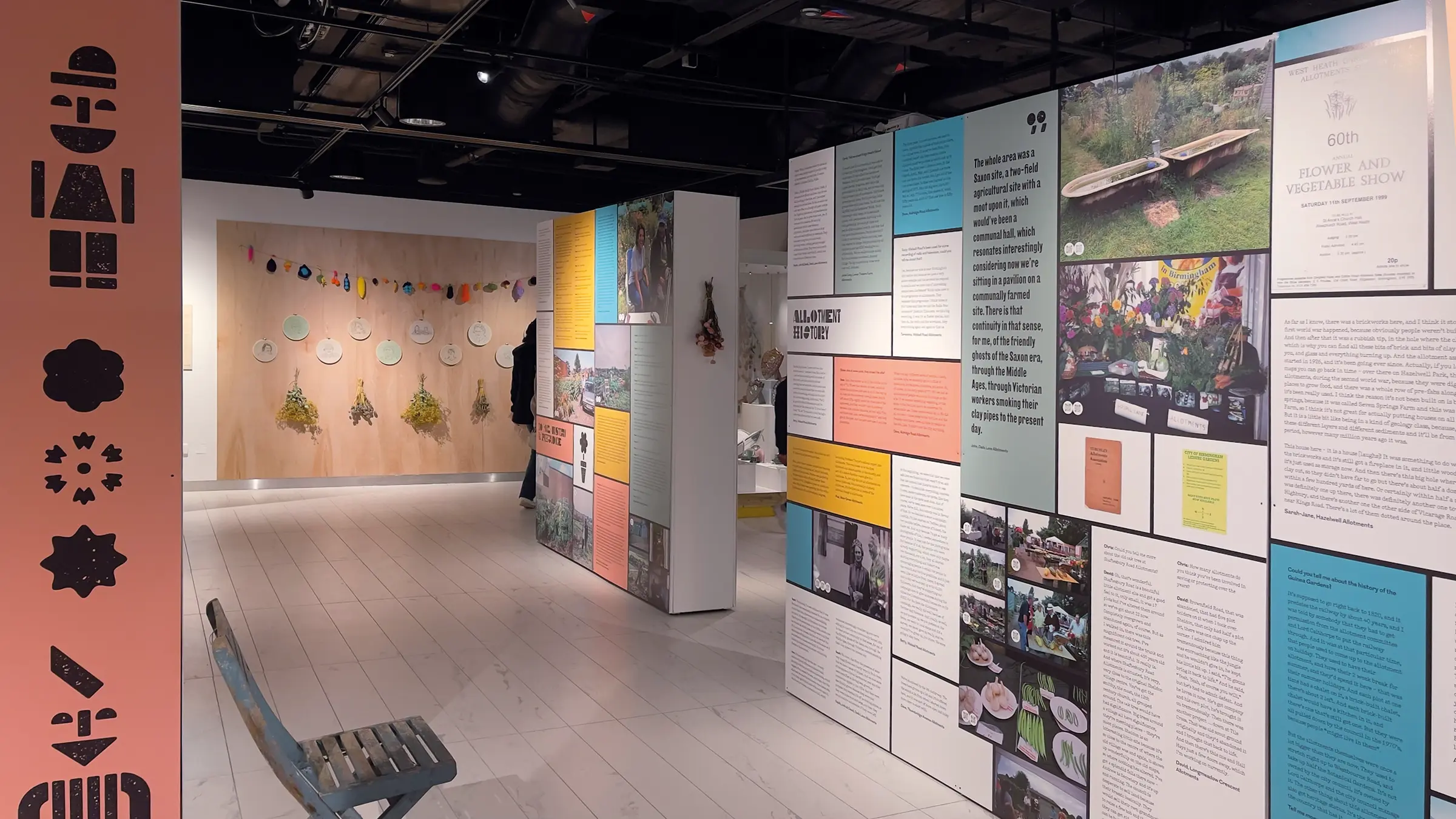 An image of multicoloured exhiition panels in a white gallery space. To the left is a pink panel, with illustrations of people and flowers made from a woodblock typeface.