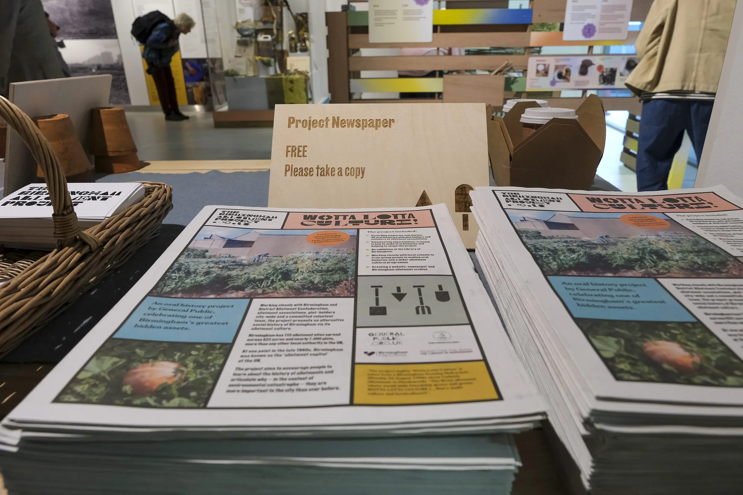 An image of a pile of newspapers on a gallery table. The cover of the papers has a grid of images and text, with multicolored panels and the headline 'WOTTA LOTTA CULTURE!'. A sign in the background reads 'Project Newspaper. Free. Please take a copy'
