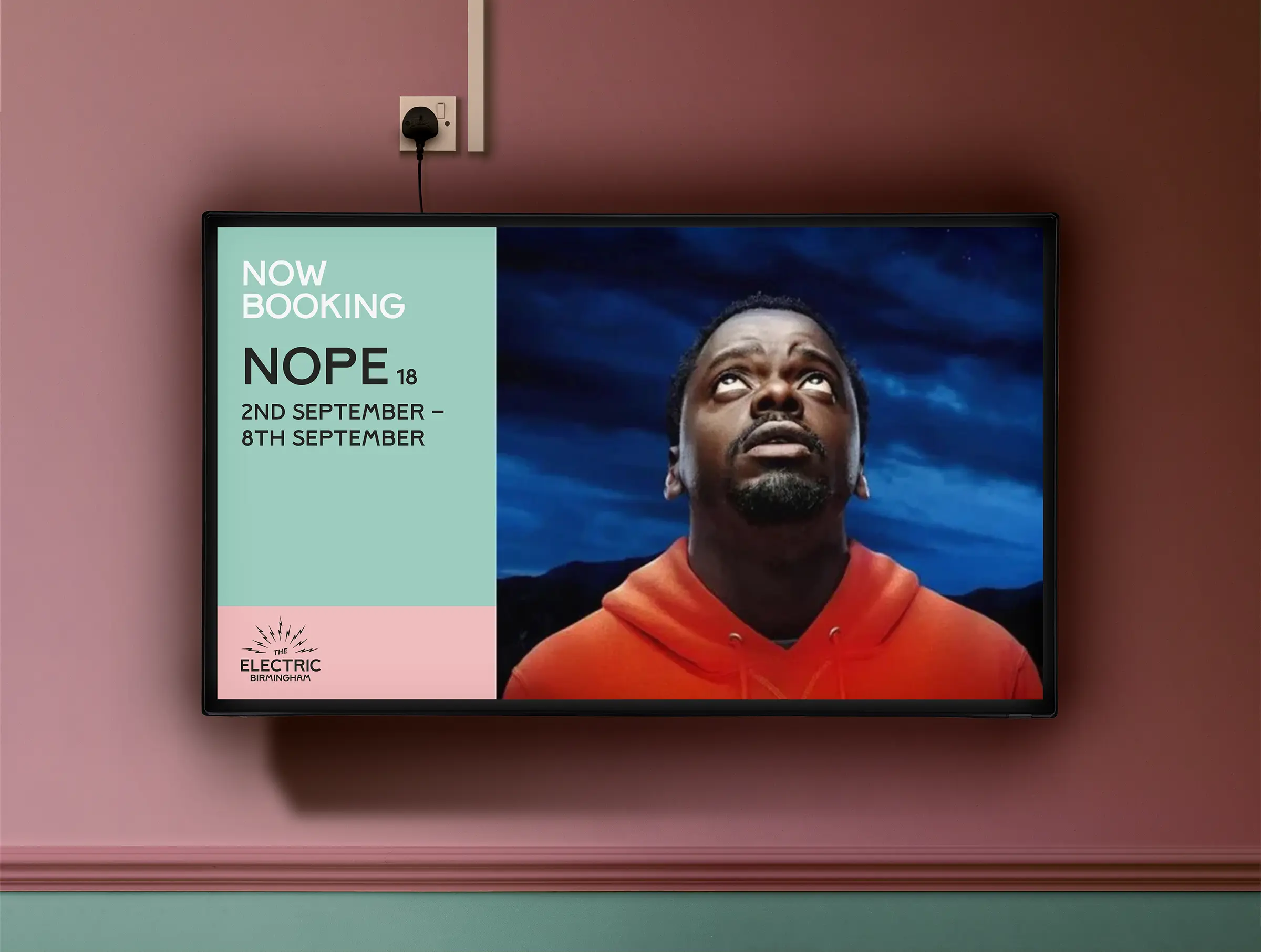 An image of a TV screen against a pale pink wall in the foyer of The Electric. An image of a man looking upwards to a darkened blue sky covers the right 2/3 of the screen, with a coloured panel on the left and text reading 'Now Booking, Nope (18), 2nd September – 8th September.
