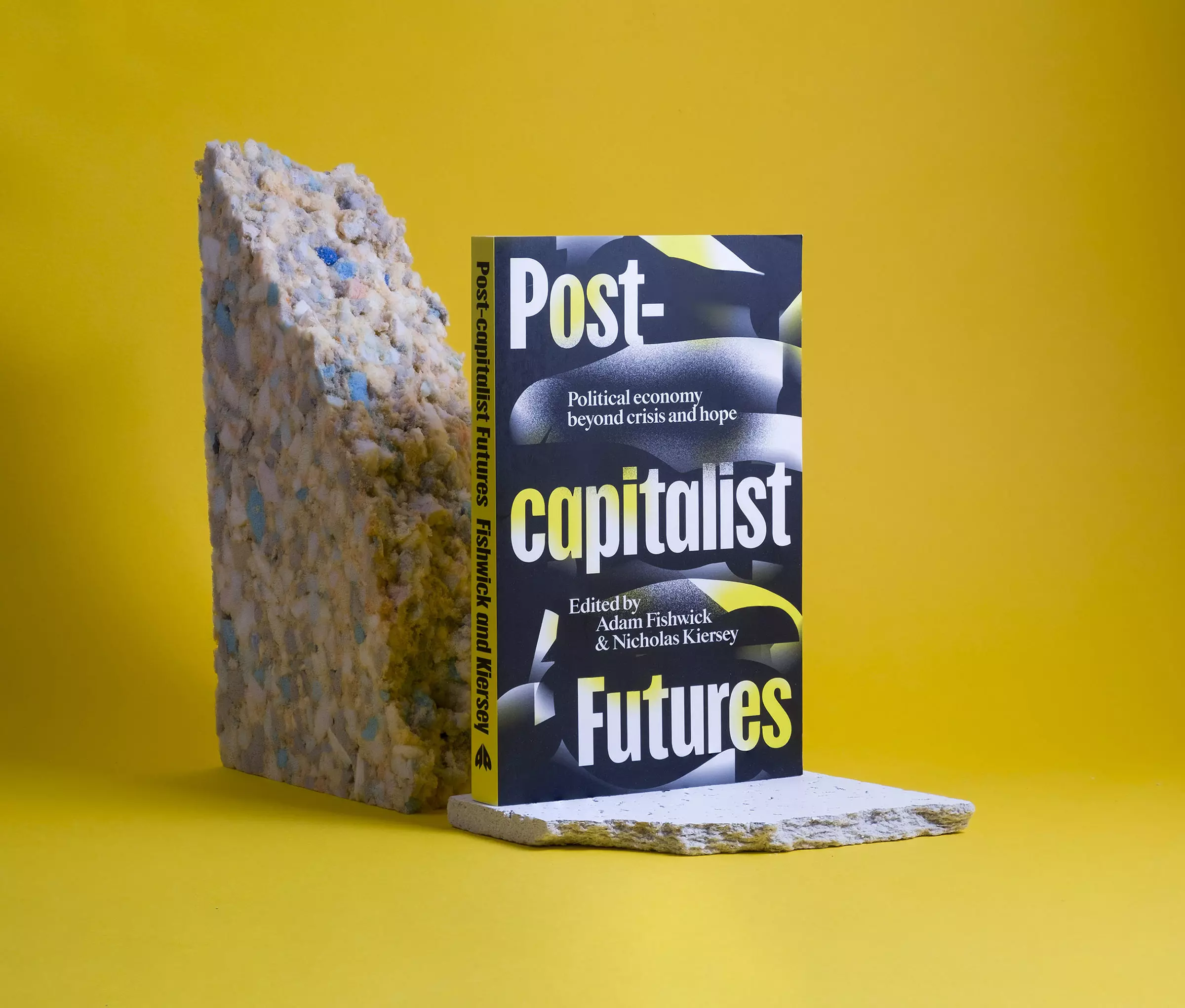 A yellow and black book cover stands on a broken ceiling tile in front of a roughly torn piece of packing foam, all agaisnt a yellow background. The cover features irregular shapes and text reading 'Post-capitalist Futures.