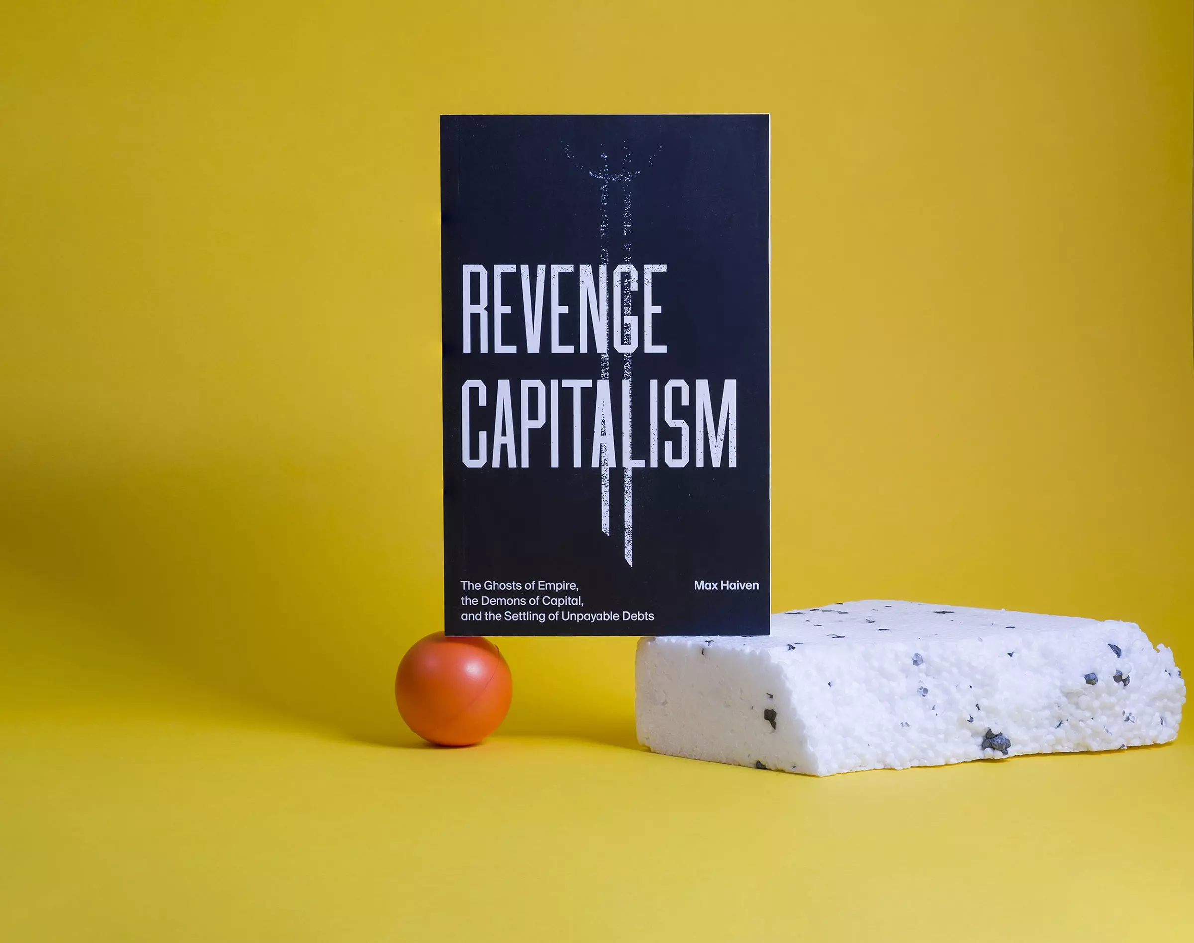 A black book balances on a slab of polystyrene and a small, orange ball in front of a yellow background. The white cover text reads 'Revenge Capitalism'.