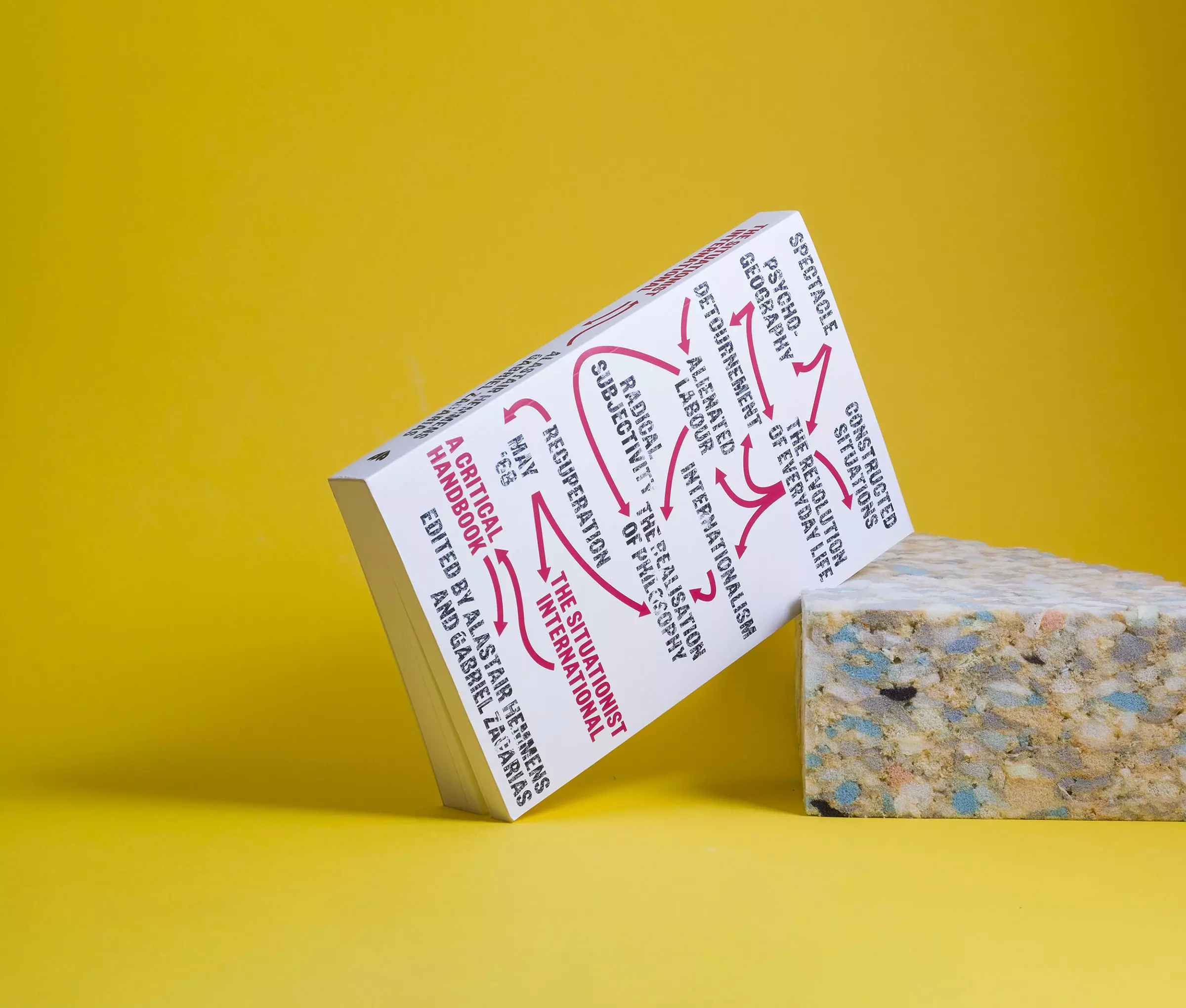 A white book leans against a piece of roughly torn packing foam against a yellow background. The cover features a graphic based on Guy Debord's 'The Naked City', with Situationist concepts in black text connected by red arrows.