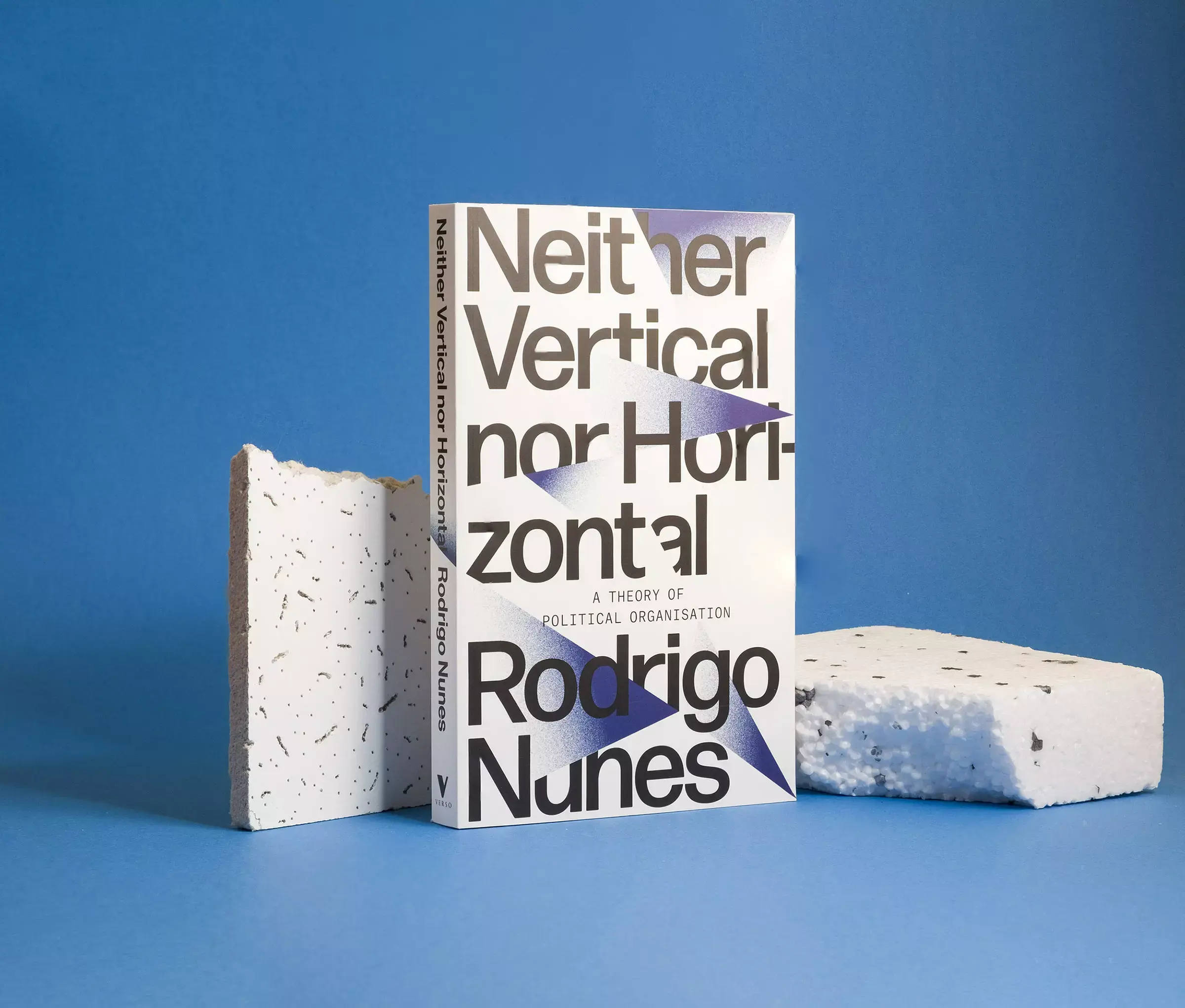 A white book cover with gradient triangles and broken text reading 'Neither Vertical Nor Horizonal', standing amongst broken materials against a light blue background.