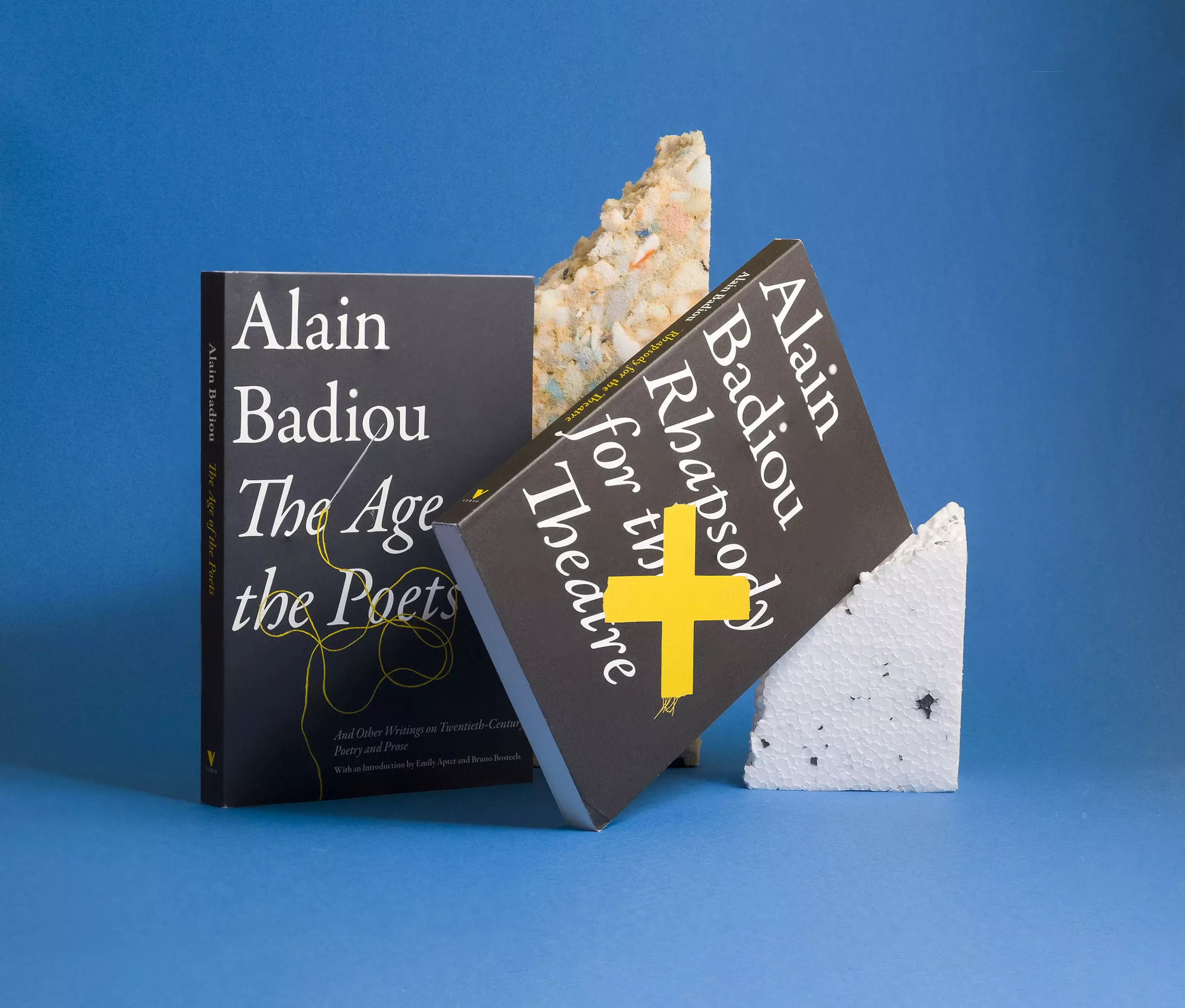Two black book covers with white serif text reading 'Rhapsody for the Theatre' and 'The Age of the Poets'. The Age of the Poets is standing, and has a yellow thread overlaying the text, with a torn piece of packing foam visible behind it. Rhapsody for the Theatre is leaning at a 45 degree angle against a piece of polystyrene, and has a yellow cross made of tape overlaying the type. Both are photographed against a light blue background.