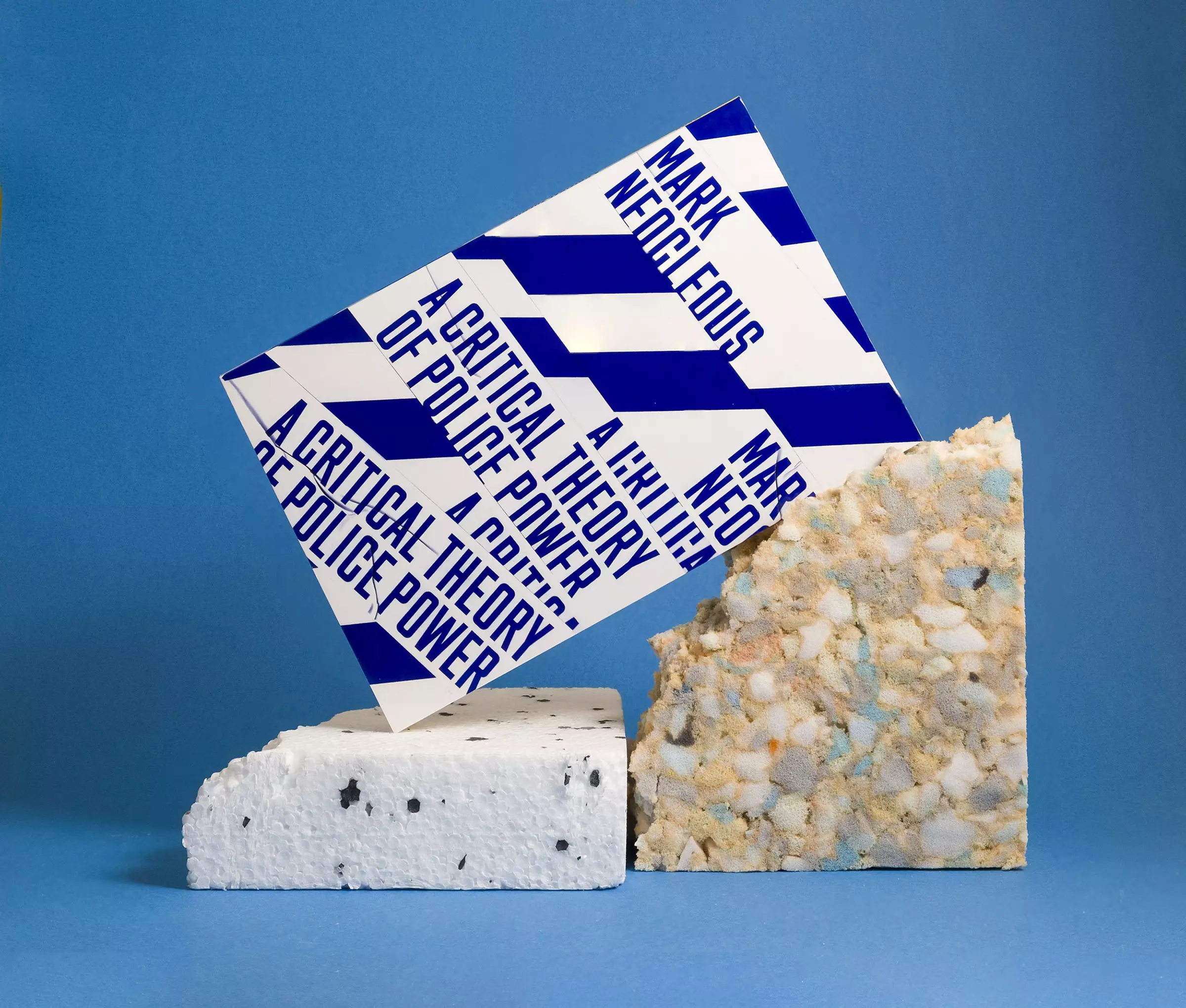 A book cover with a trompe l'oeil effect of being wrapped in police tape reading 'A Critical Theory of Police Power'. It stands on a thick piece of polystyrene and leans against a roughly torn piece of packing foam, all photographed against a light blue background.