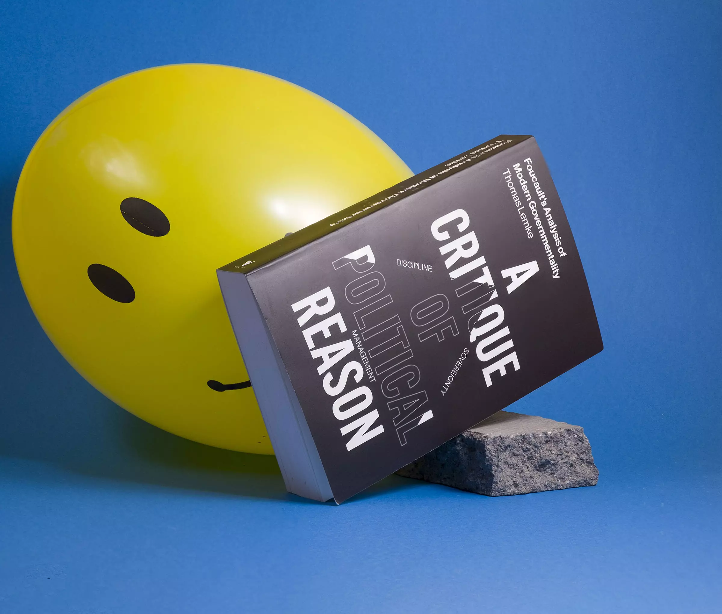 A black book cover reading 'A Critique of Political Reason', leaning against a small piece of paving slab against a light blue background. Behind the book, a large yellow smiley face balloon peeks out. 