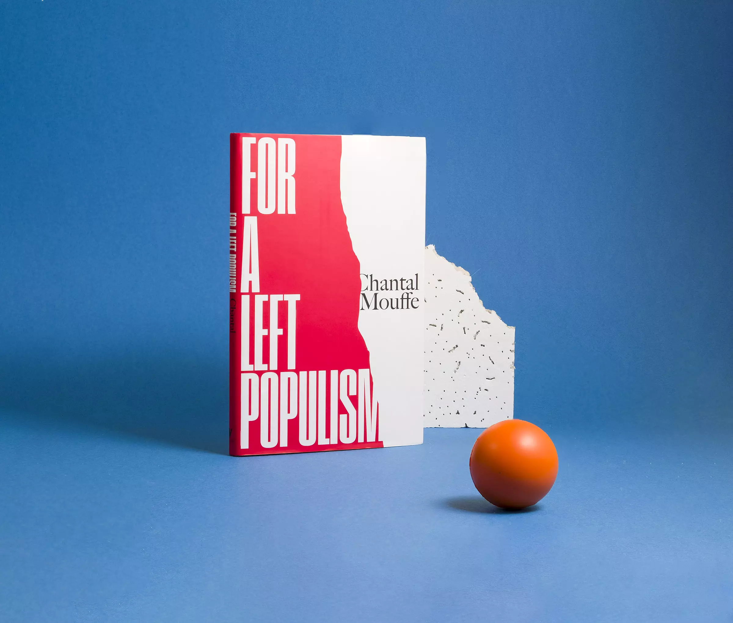 A book cover with a torn red shape to the left, and text reading 'For a Left Populism', standing in front of a broken ceiling tile against a light blue background. A small orange ball sits in the foreground.