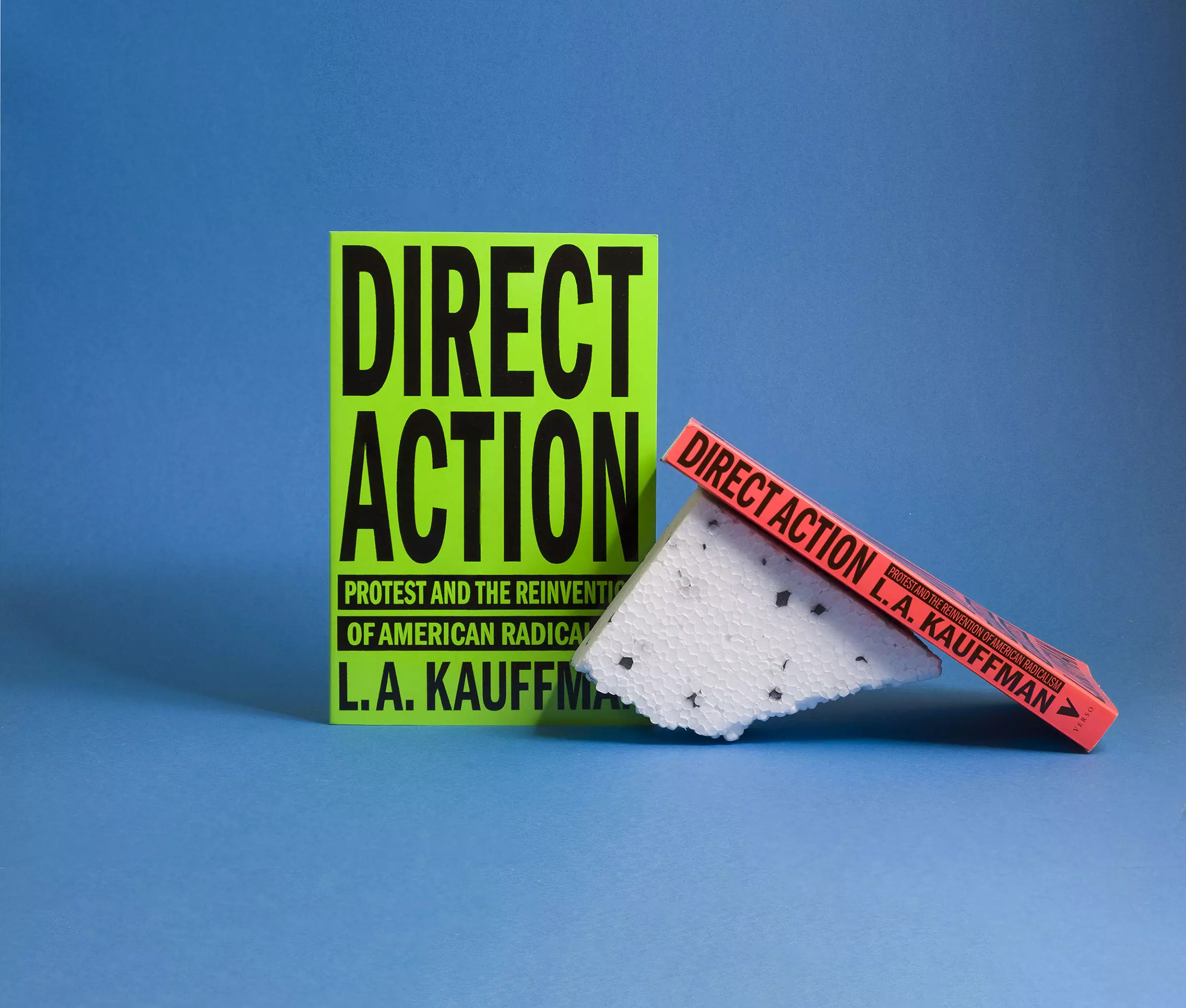 Two book covers, one fluoro green standing up square to the camera, the other fluoro red leaning at angle on a piece of broken polystyrene with only the spine visible. Both have text reading 'Direct Action', and are photographed against a light blue background.