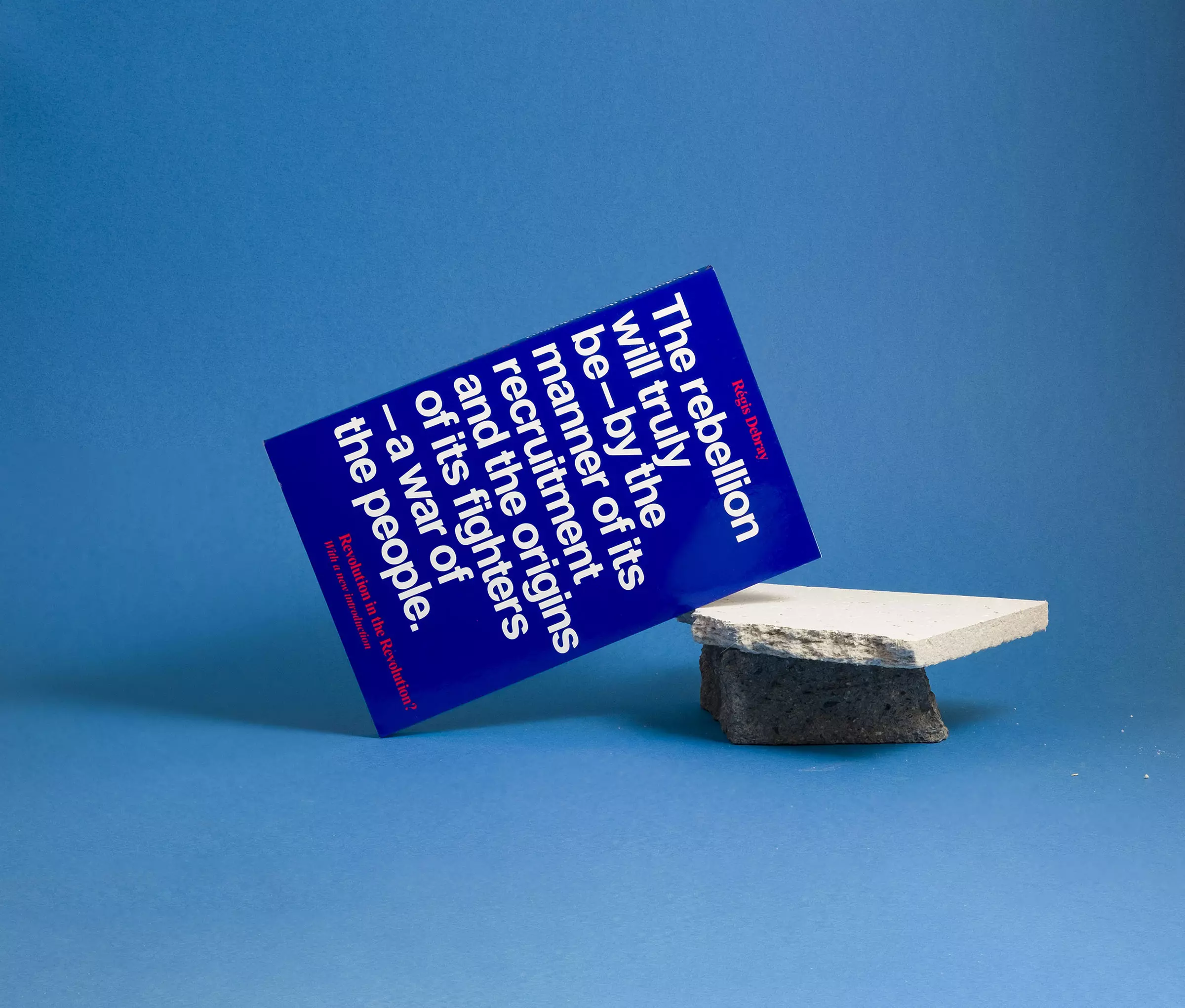 A blue book cover with white text reading 'The rebellion will tryly be – by the manner of its recruitment and the origins of its fighters – a war of the people.' It's leaning against a piece of broken polystyrene sitting on top of a small piece of paving slab against a light blue background.
