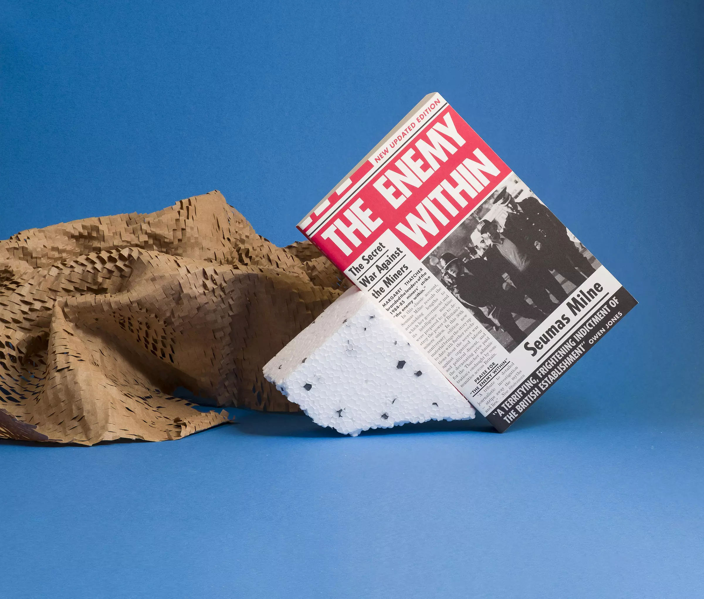A red and white book cover, styled to look like a 1980s edition of a tabloid newspaper, with a black and white photograph of Arthur Scargill and headline text reading 'The Enemy Within'. It leans against a small block of polystyrene against a light blue background, with brown perforated packing paper sitting behind the book.