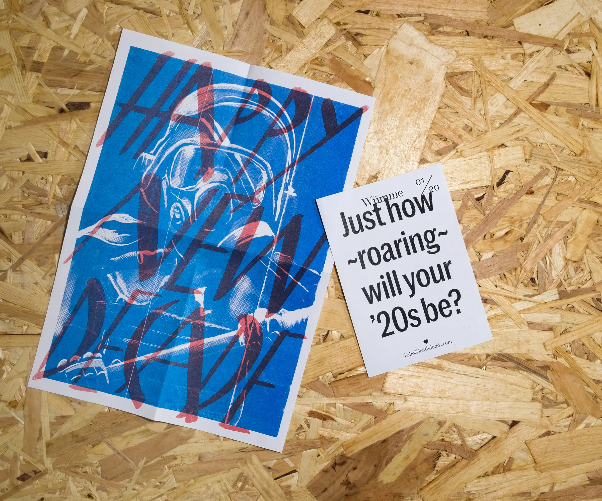 An A4, blue drawing of a riot police officer overlaid with red, hand-drawn text reading 'Happy New Decade'. A smaller, A6 piece of paper reads 'Just how roaring will your 20s be?'. Both sit on a background of OSB.