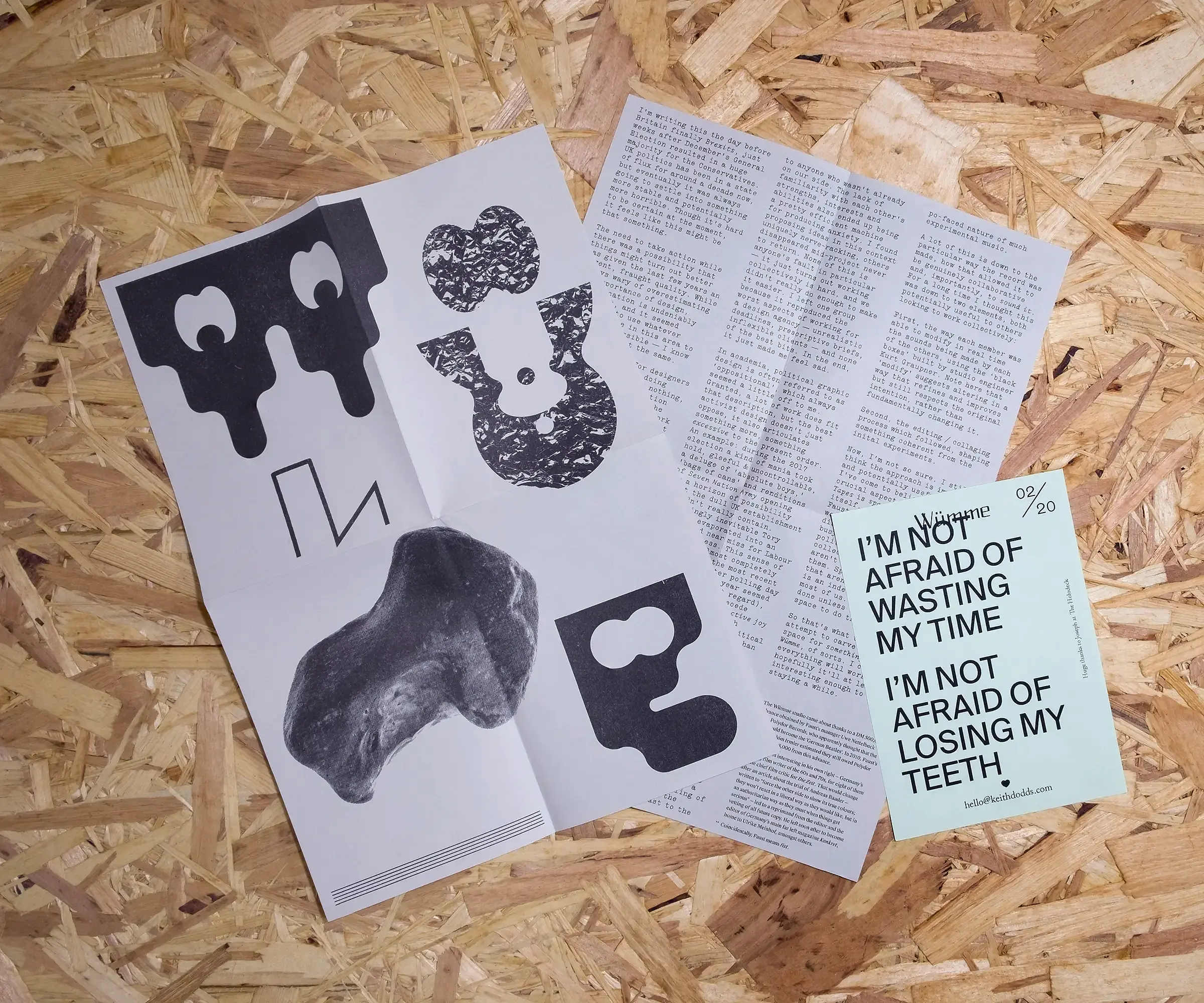 An A4, folded piece of paper with a type collage reading 'WÜMME' on one side, and densely typeset text on the other. A smaller, A6 piece of blue paper reads 'I'm not afraid of wasting my time. I'm not afraid of losing my teeth'. Both sit on a background of OSB.