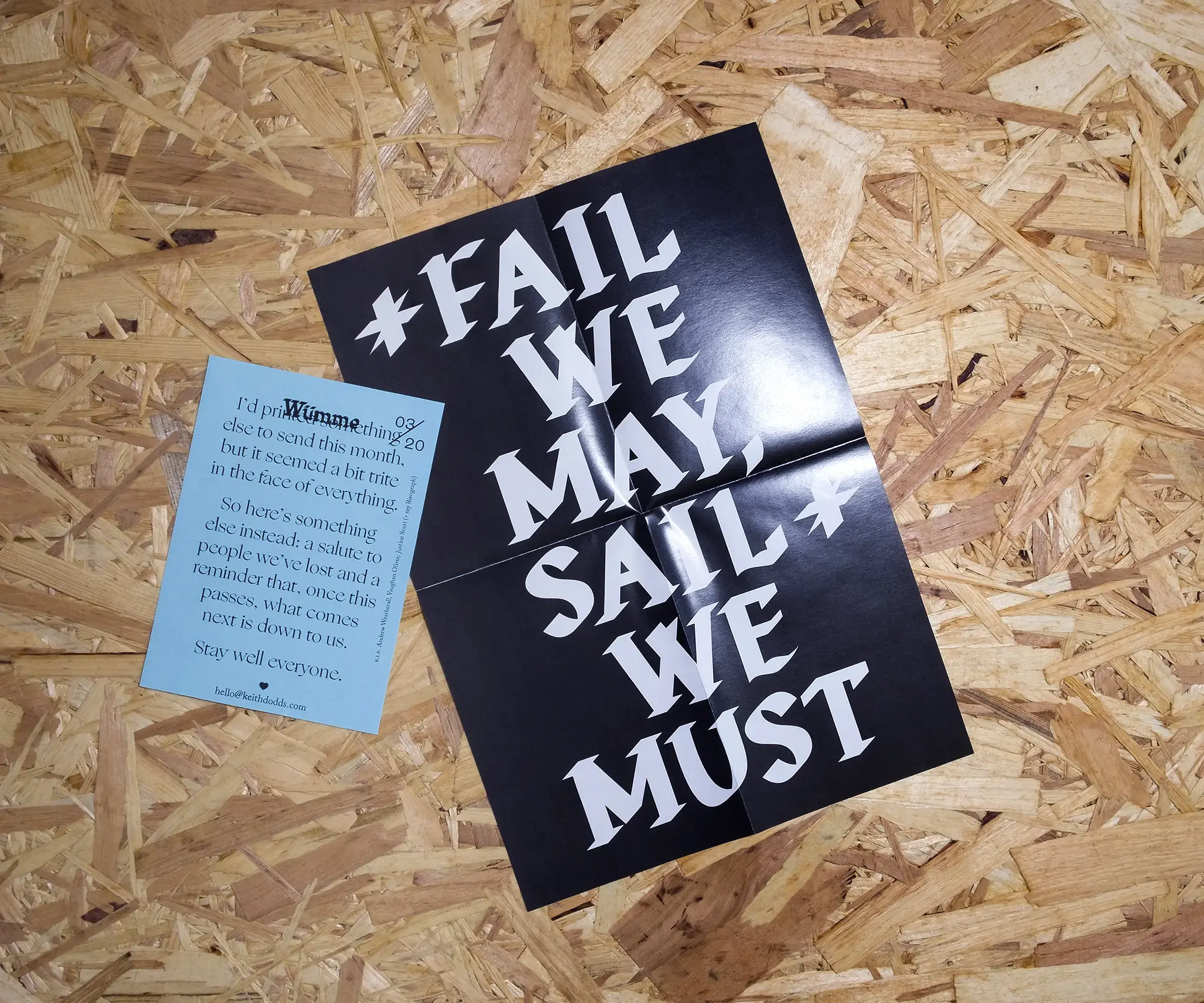 An A4, folded piece of black paper reading 'FAIL WE MAY. SAIL WE MUST'. A smaller, A6 piece of blue paper with a dense paragraph of text. Both sit on a background of OSB.