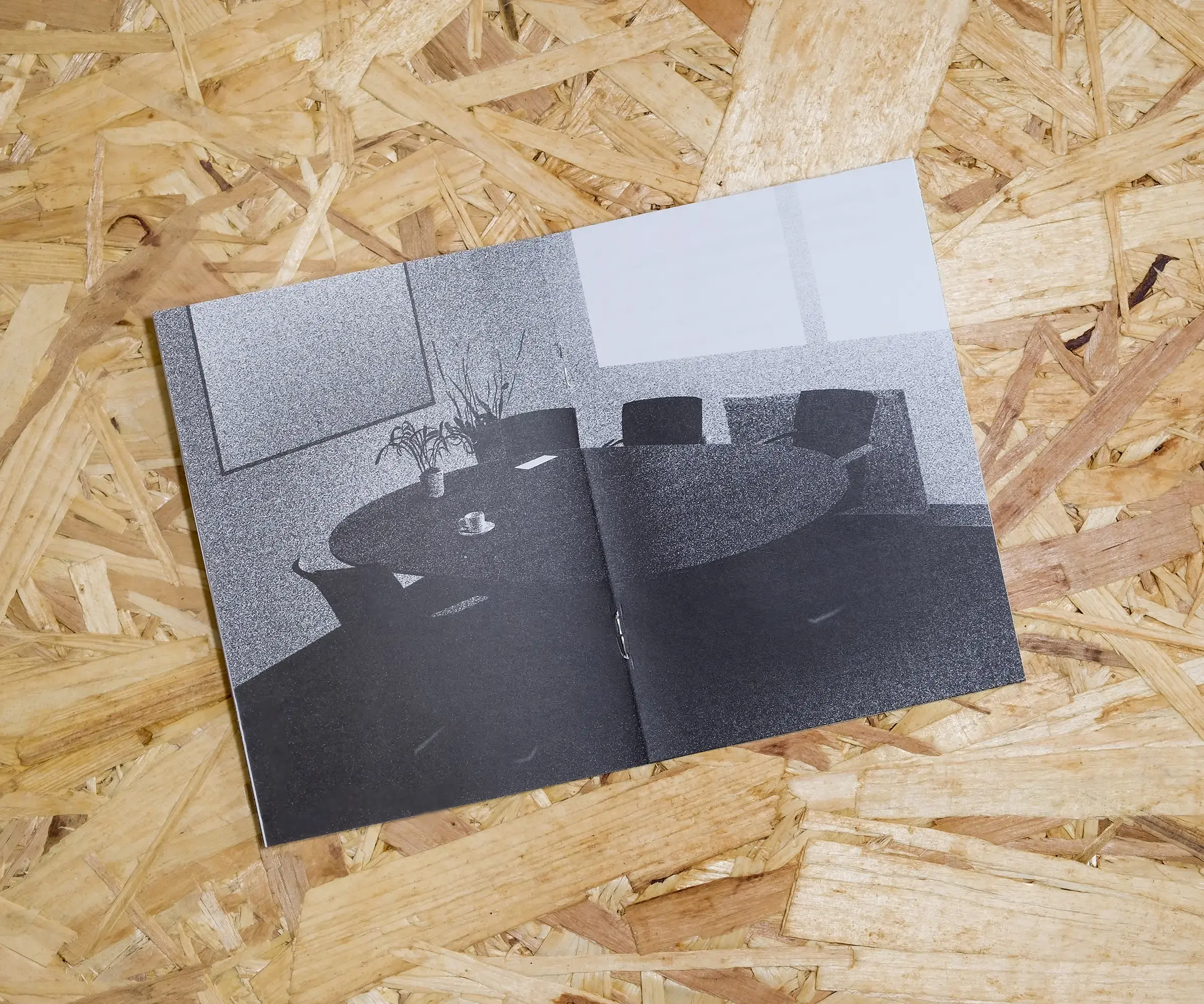 An open A6 booklet, with white pages and a grainy, realistic illustration of a boring office meeting table, on a background of OSB.