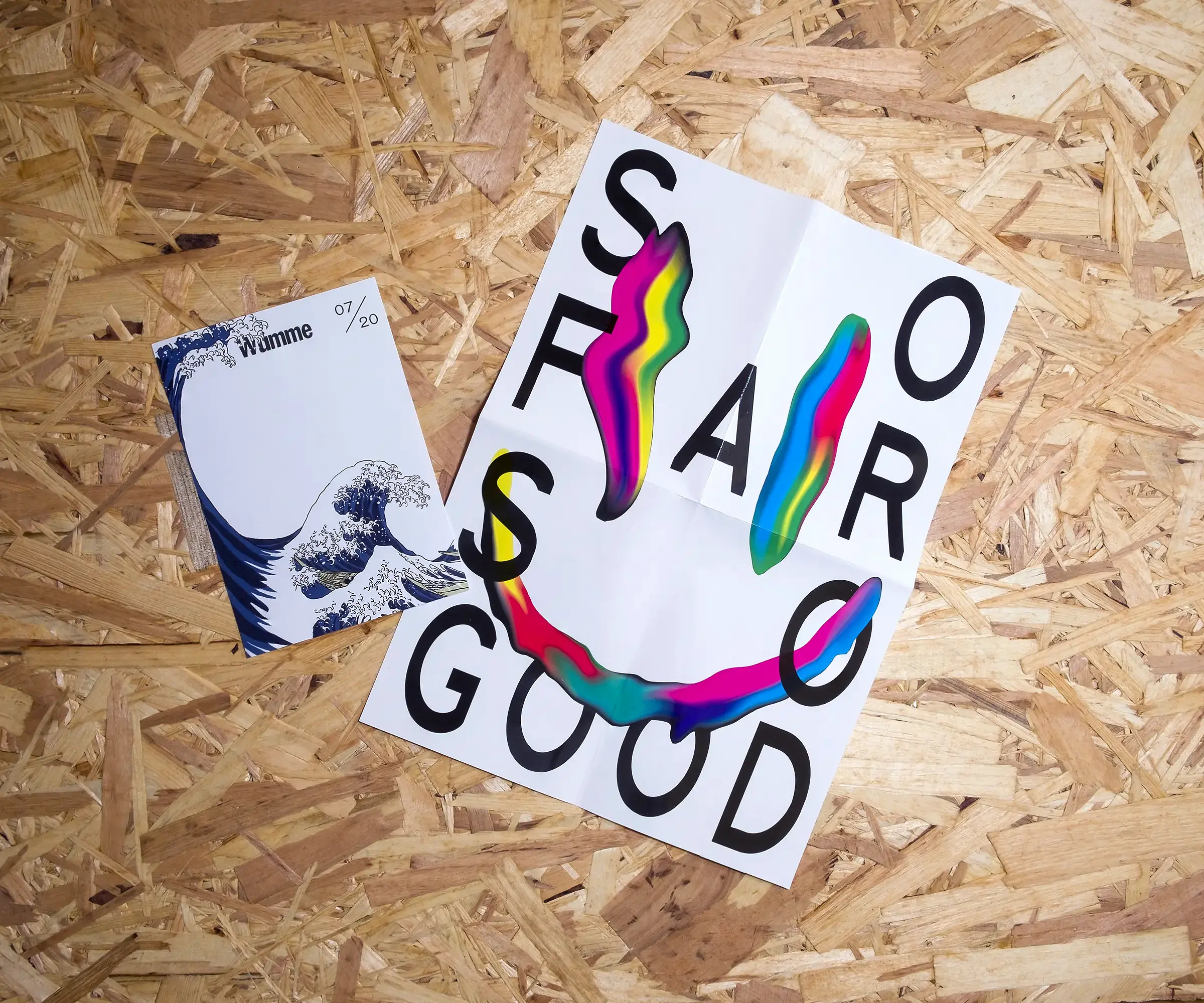 A white, A6 piece of paper features a colour illustration of Hokusai's 'The Great Wave', with a much bigger wave appearing to follow behind it. Beside it is a folded, white A4 piece of paper with a multicoloured, distorted smiley face, and the text 'SO FAR SO GOOD'. Both sit on a background of OSB.