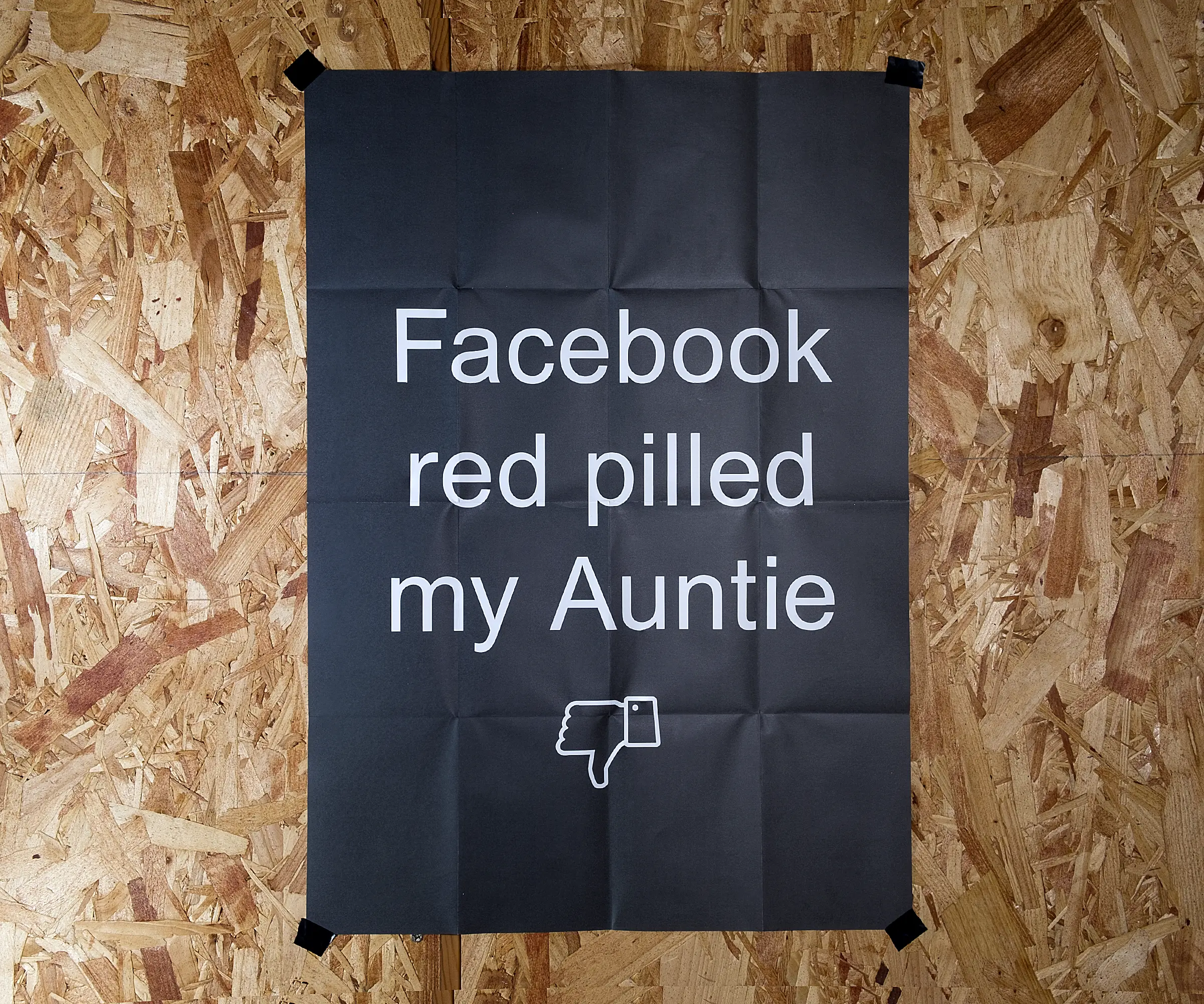 A black, A2 poster on a background of OSB, with the words 'Facebook red pilled my Auntie' and a thumbs down symbol.