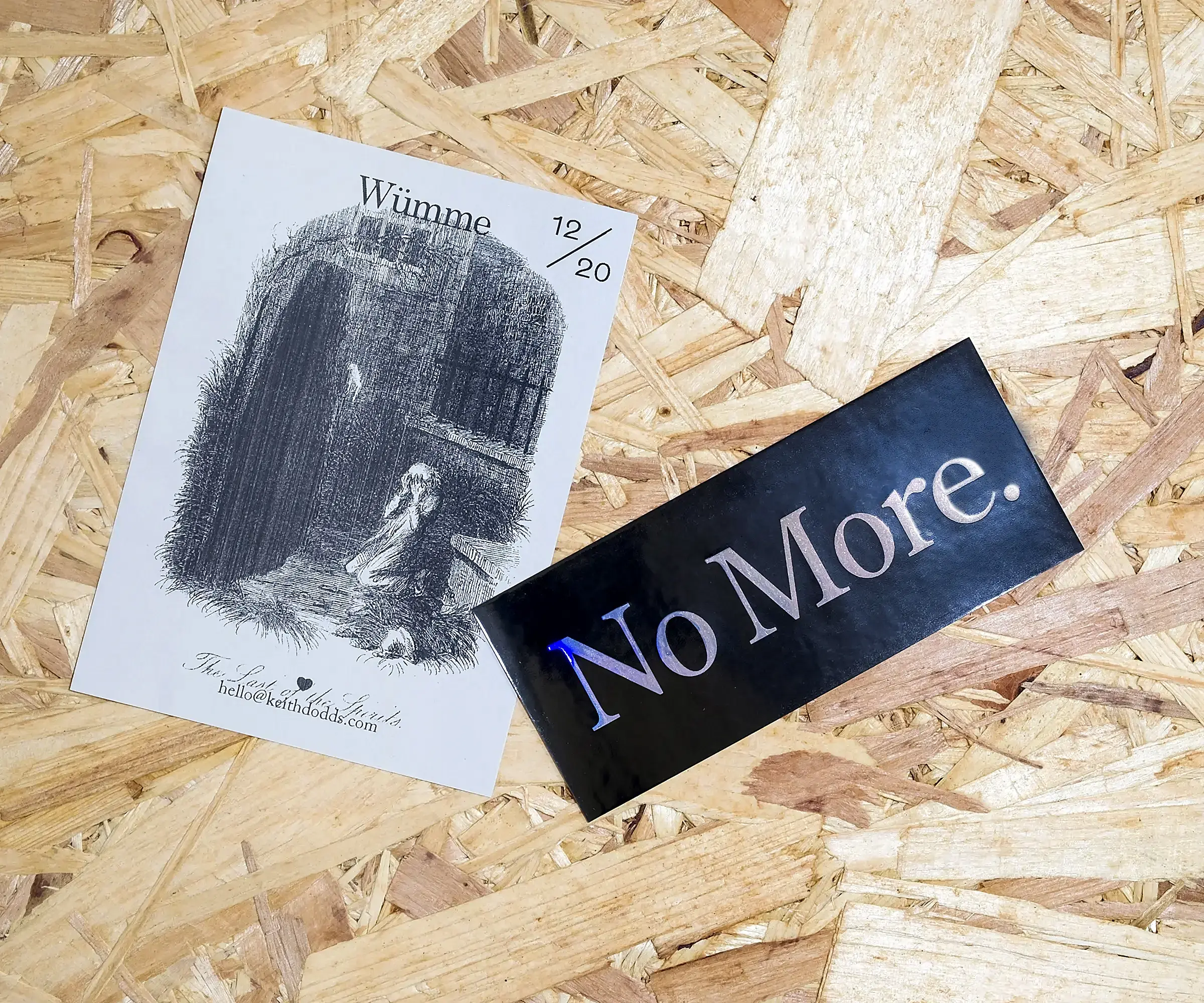 An A6, grey piece of paper featuring an illustration of The Ghost of Christmas Yet to Come from Charles Dickens' 'A Christmas Carol'. Beside it is a small sticker, with a black background and holographic text, reading 'No More.'
